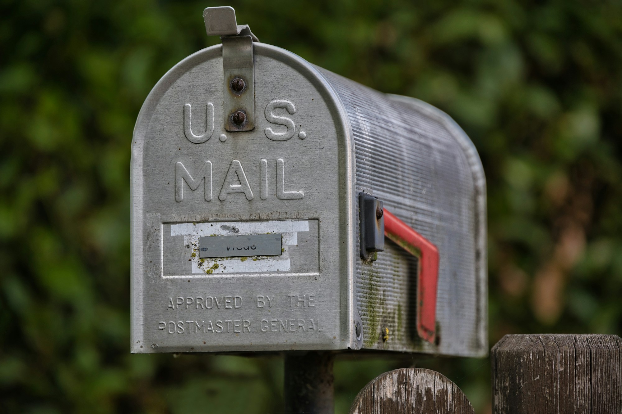 Briefkasten aus gebürstetem Metall mit aufgestellter roter Fahne vor einer unscharfen grünen Hecke