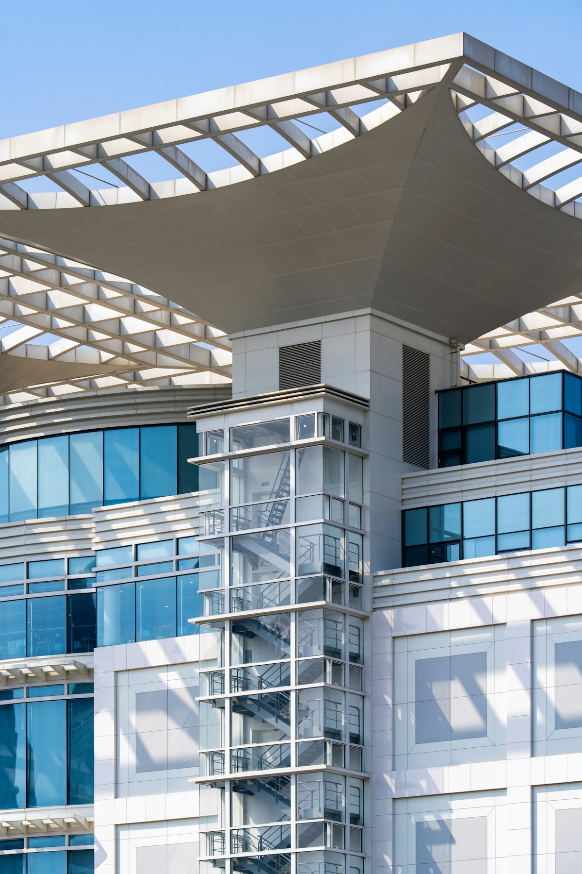 Tour d’escalier vitrée d’un immeuble de bureaux moderne sous une vaste toiture ajourée, ciel bleu