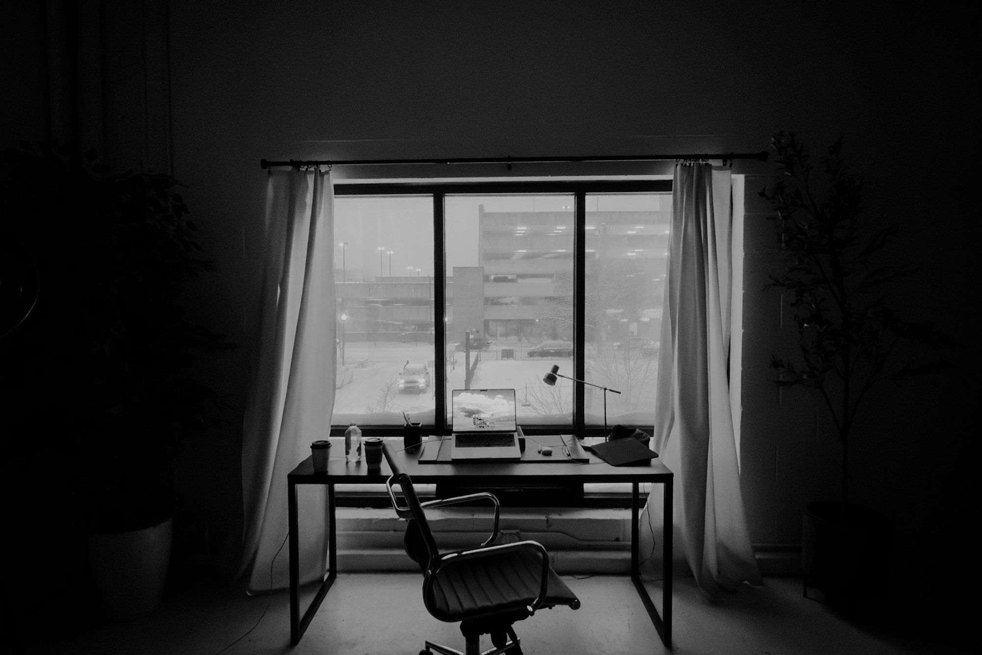 Black-and-white photo of an empty office chair in front of a desk with a laptop and lamp, facing a large window with white curtains overlooking a snowy street