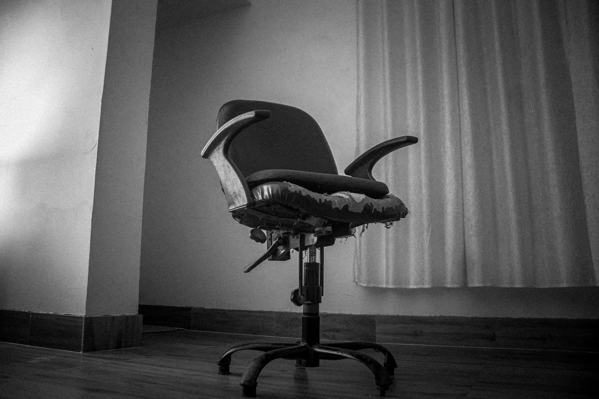 A worn, empty office chair in front of a curtained window, in black and white