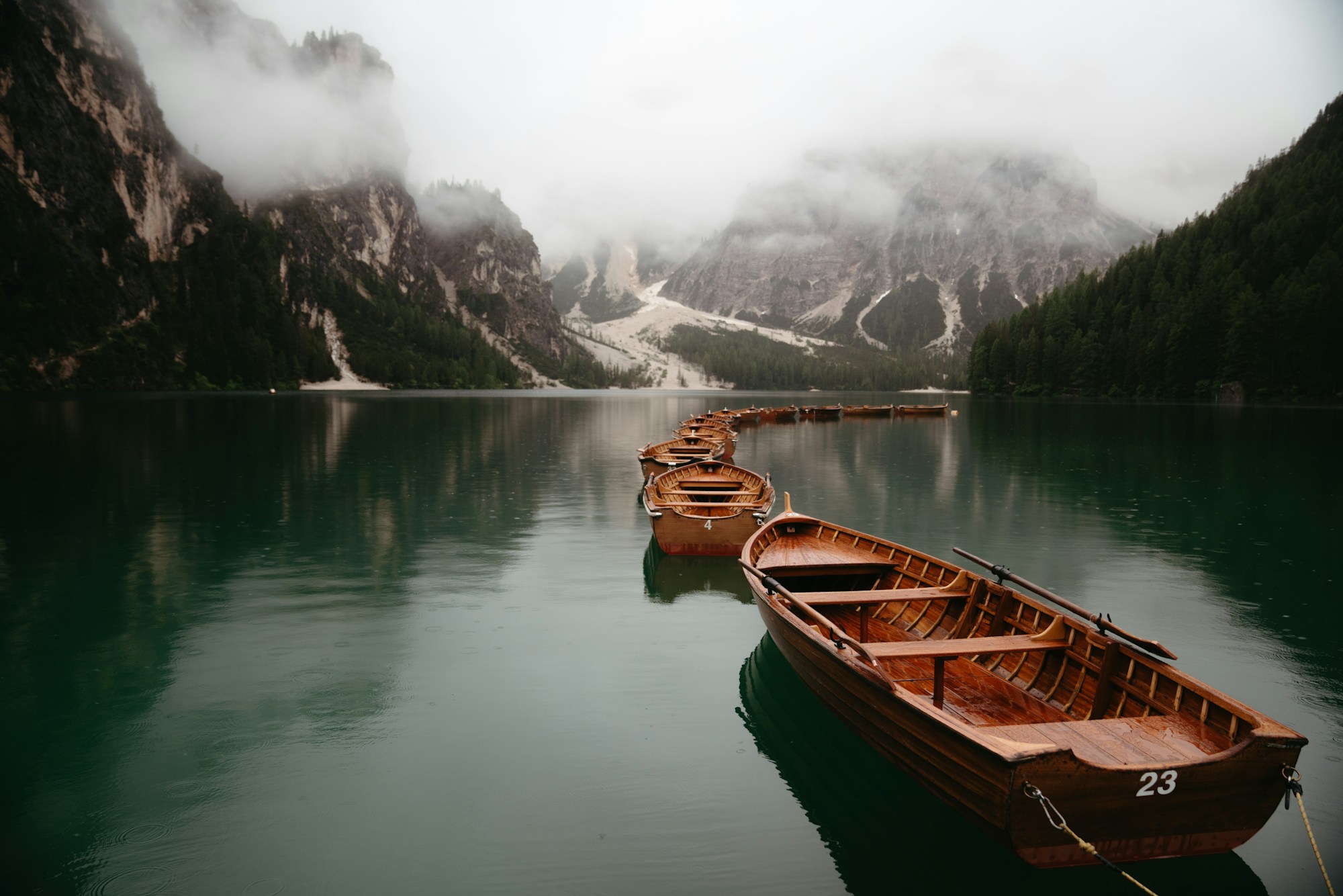 File de barques en bois amarrées sur un lac alpin aux eaux vertes, sous des parois rocheuses prises dans la brume