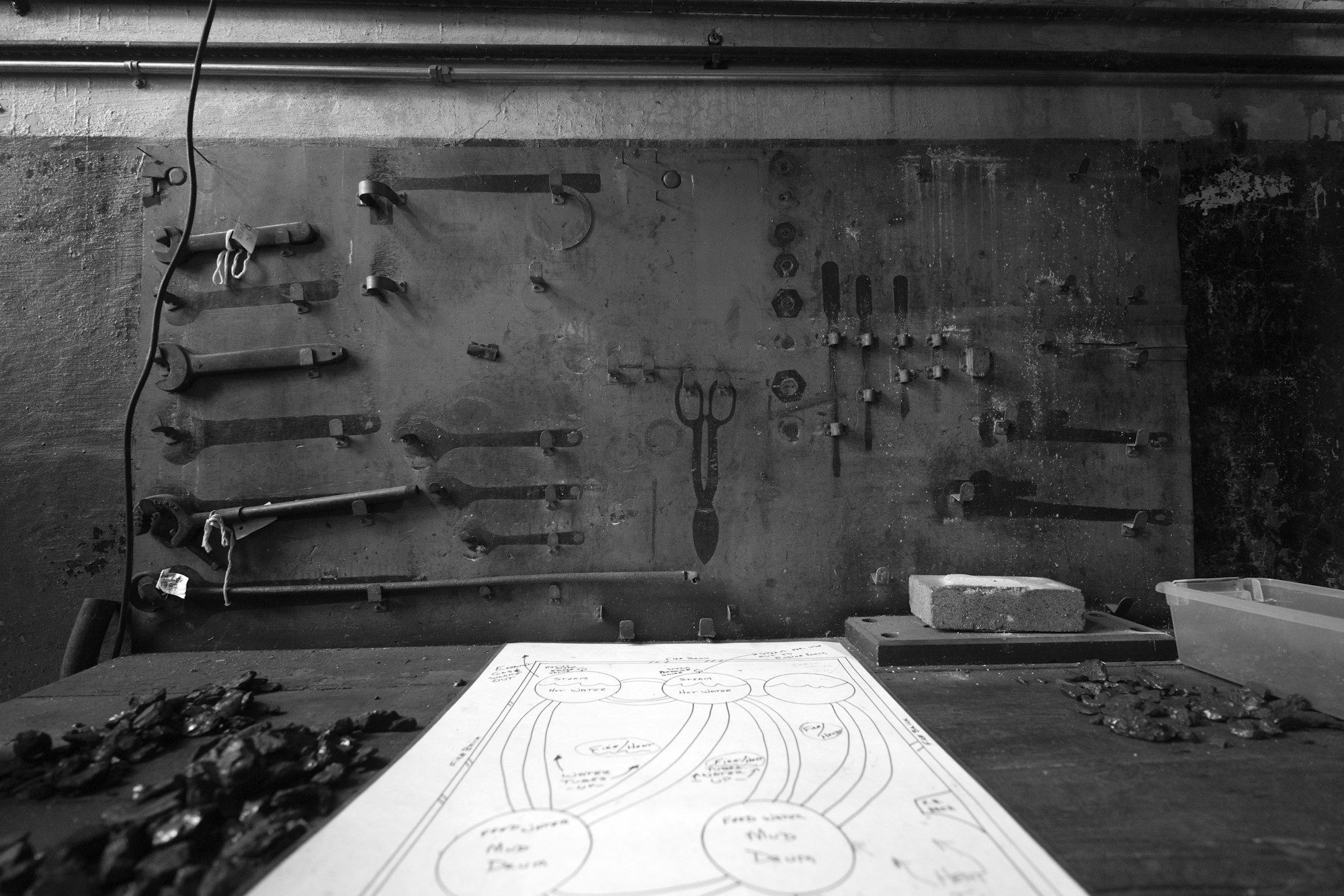 Mur d'atelier en noir et blanc, outils suspendus à des crochets, plusieurs emplacements vides, un schéma dessiné à la main posé sur l'établi