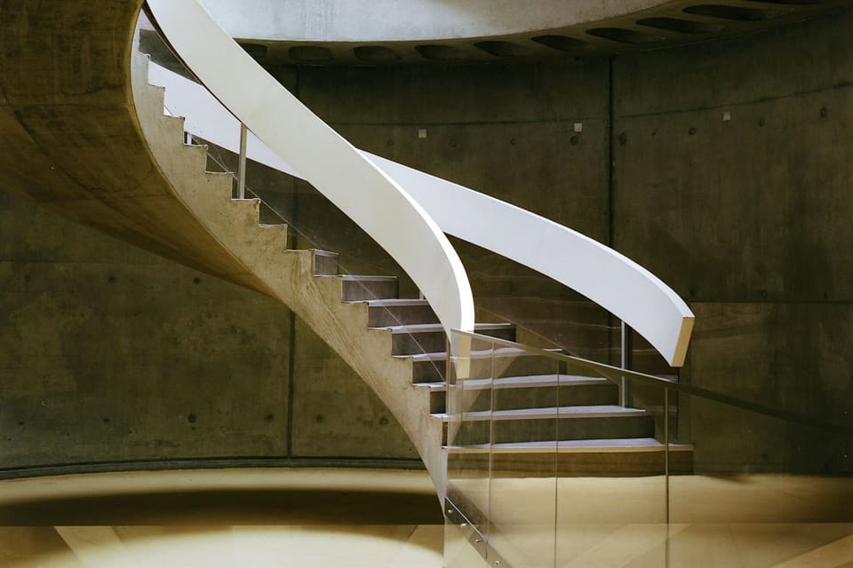 Geschwungene Betontreppe mit weissem Geländer, die zu einem runden Oberlicht hinaufführt