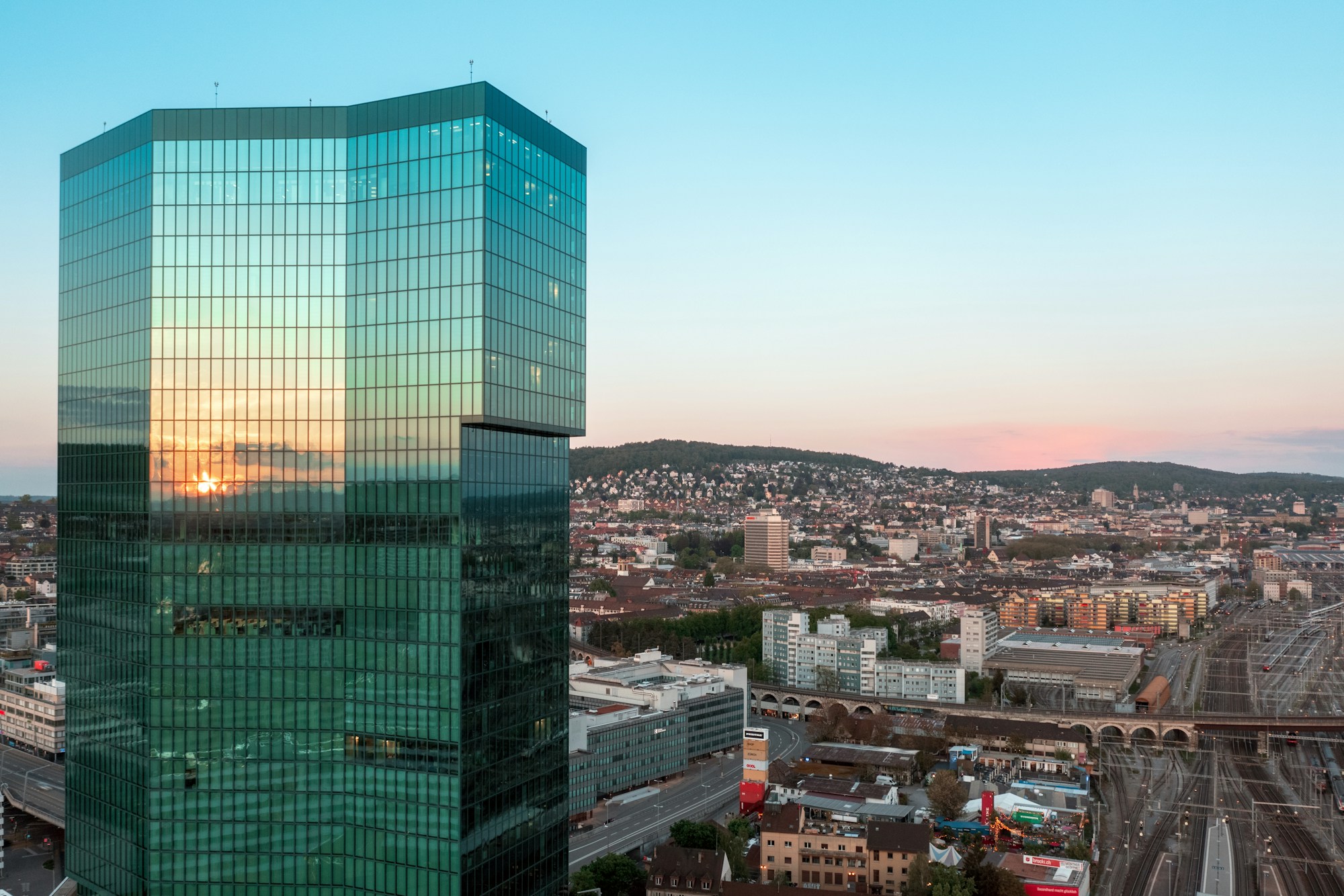 Glass office tower overlooking a Swiss city at sunrise, facade reflecting the sky