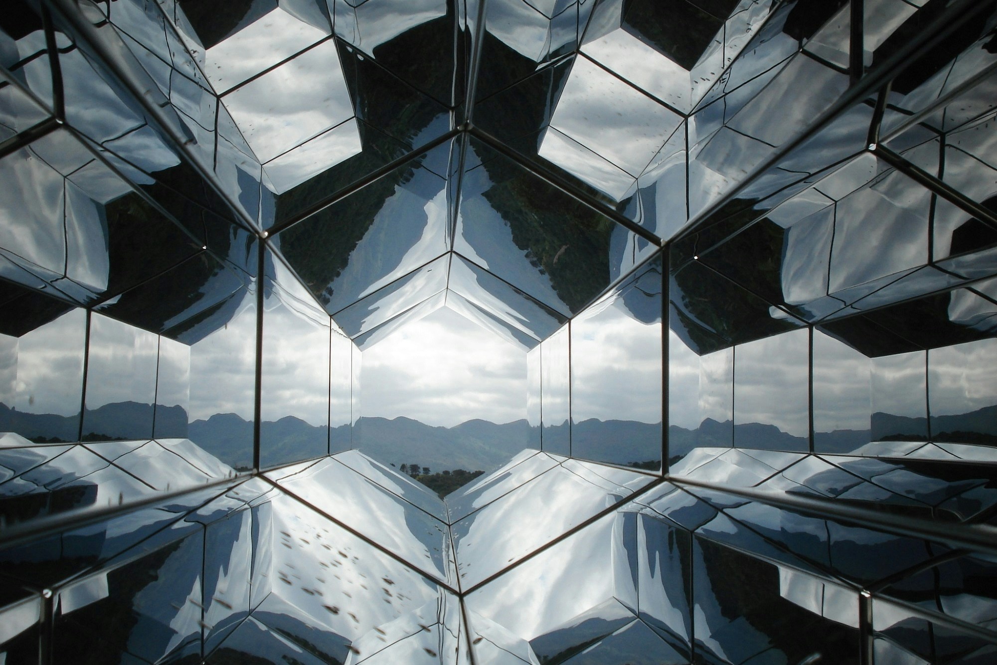 Kaleidoskopartige Struktur aus spiegelnden Glasflächen von innen gesehen, in der Mitte ein bewölkter Himmel und Berge
