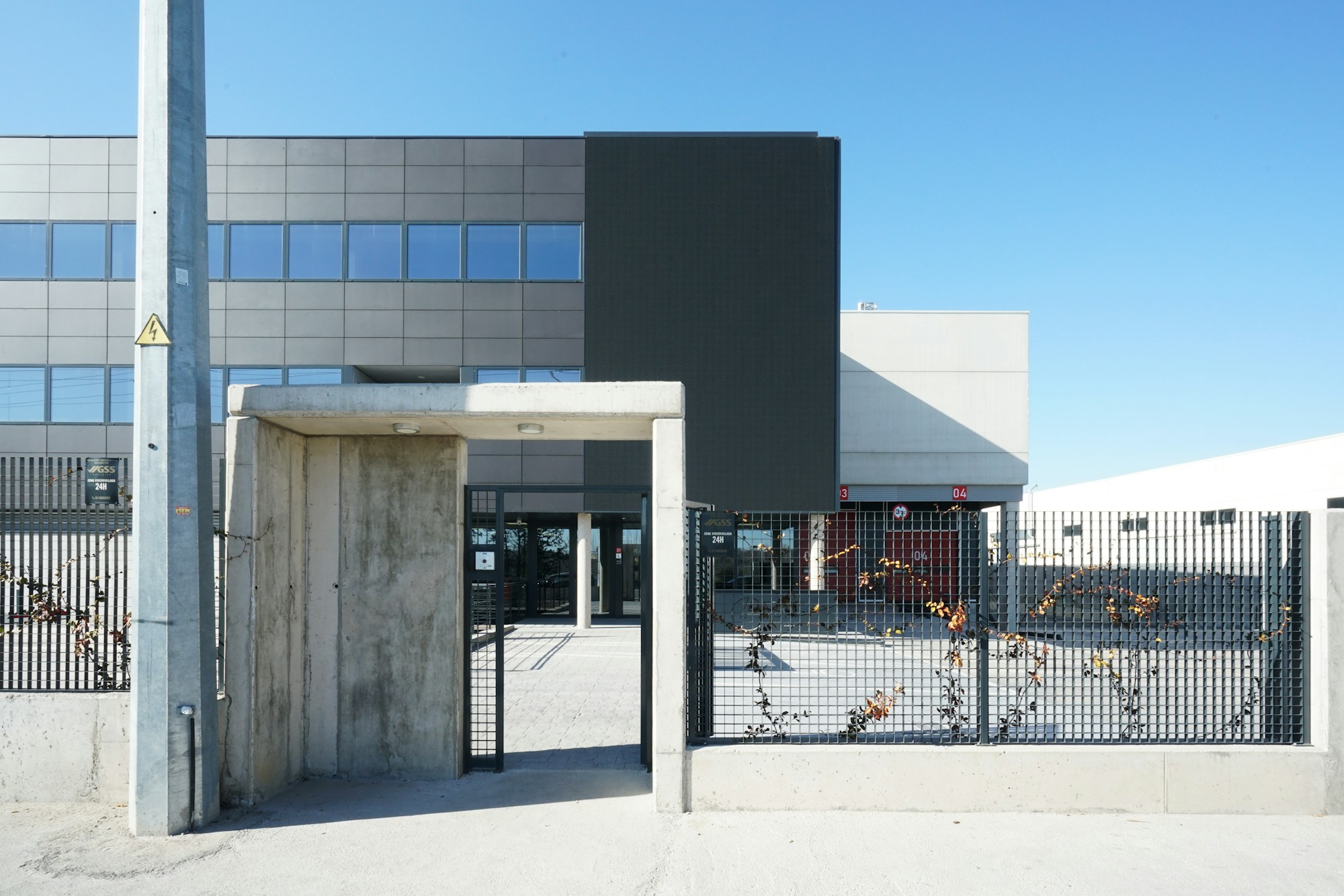 Entrée grillagée d'un bâtiment industriel au bardage gris et noir, sous un ciel bleu dégagé