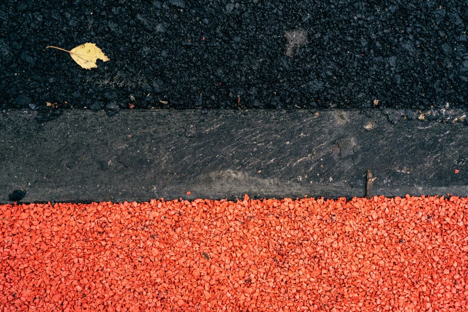 Blick von oben auf einen grauen Betonrandstein, der schwarzen Asphalt mit einem einzelnen gelben Blatt von einer Fläche aus leuchtend rotem Kies trennt