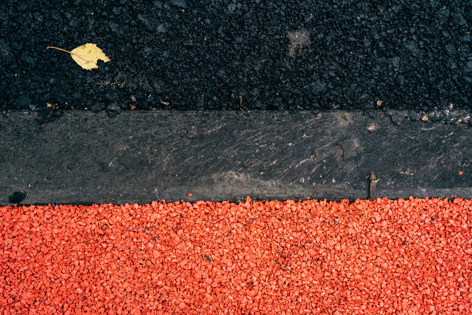 Blick von oben auf einen grauen Betonrandstein, der schwarzen Asphalt mit einem einzelnen gelben Blatt von einer Fläche aus leuchtend rotem Kies trennt