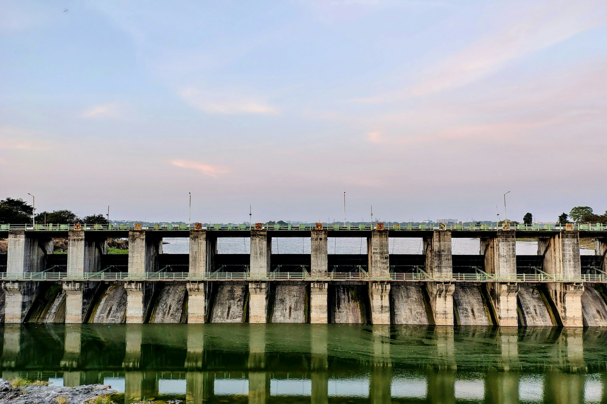 Betonwehr mit einer Reihe von Pfeilern und grünen Geländern, gespiegelt in stillem grünlichem Wasser unter einem rosa-blauen Abendhimmel