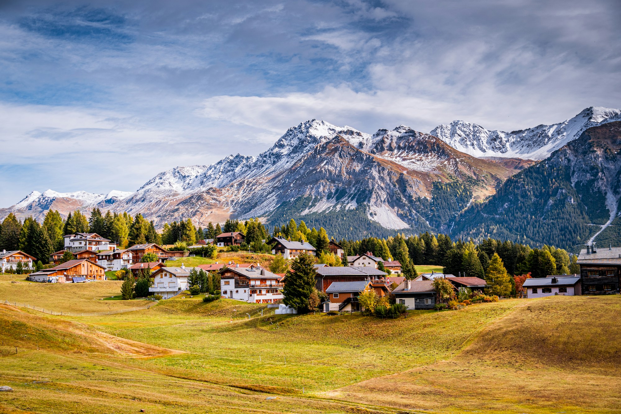Village de chalets en bois et maisons blanches sur des prairies d'automne jaunies, chaîne de montagnes enneigées et forêt de sapins à l'arrière-plan