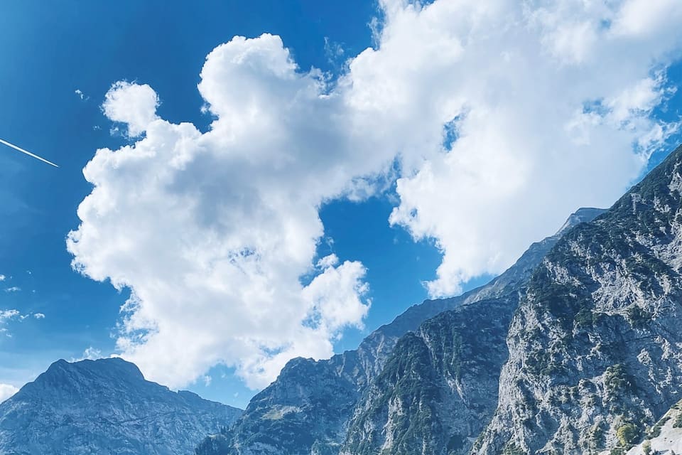 Prairie alpine verte bordée de conifères au pied de montagnes escarpées sous un ciel bleu nuageux
