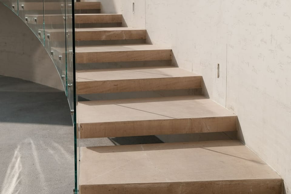 Floating stone staircase with a curved glass balustrade along a sunlit limestone wall