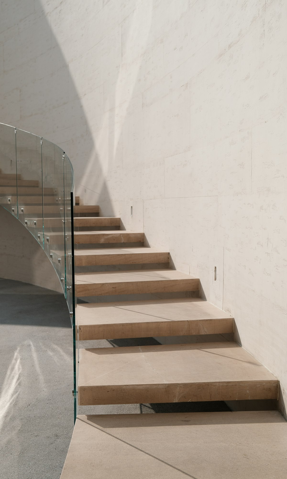 Escalier de pierre autoportant avec garde-corps en verre courbe le long d’un mur baigné de soleil