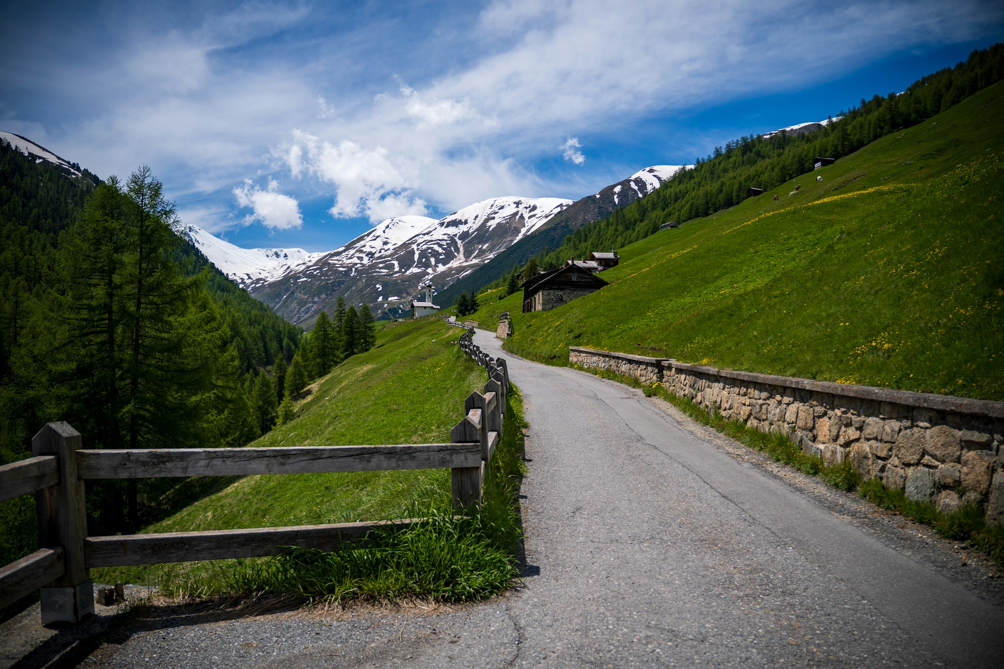 Mountain road lined with a wooden fence and a dry stone wall, chalets and a small chapel in the distance, snow-capped peaks under a blue sky