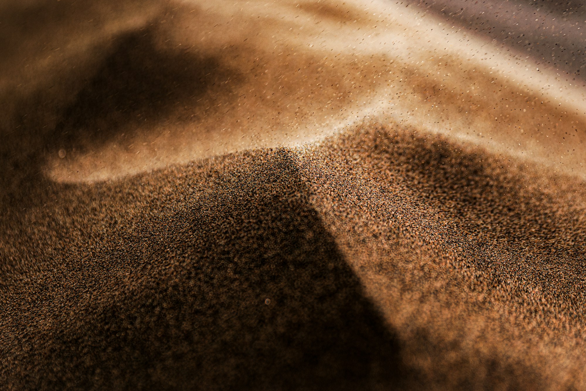 Close-up of brown sand grains forming a miniature ridge, lit from the side, with a few grains suspended in the light