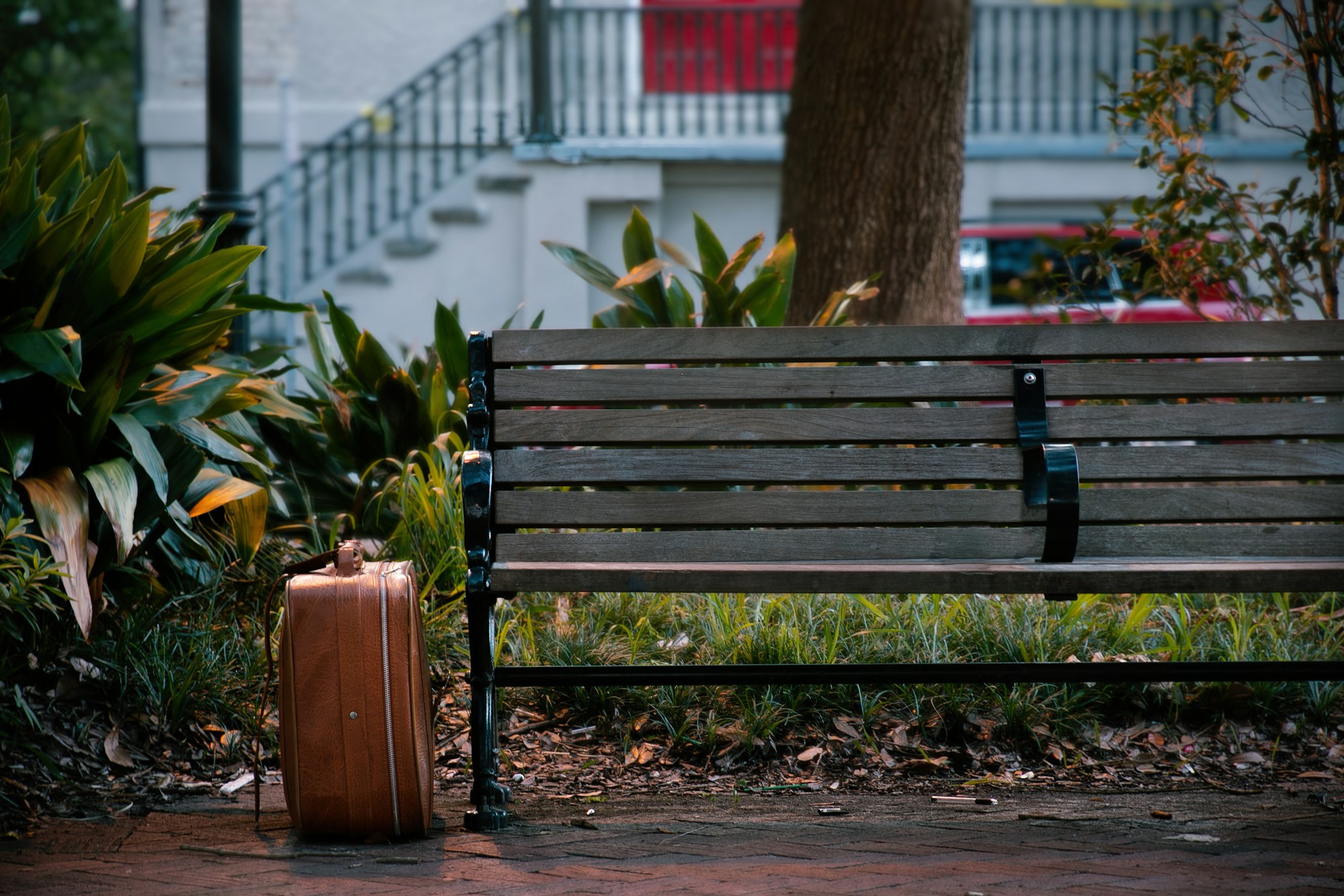Worn brown leather suitcase standing on the ground against a wooden park bench, with plants and a blurred staircase behind