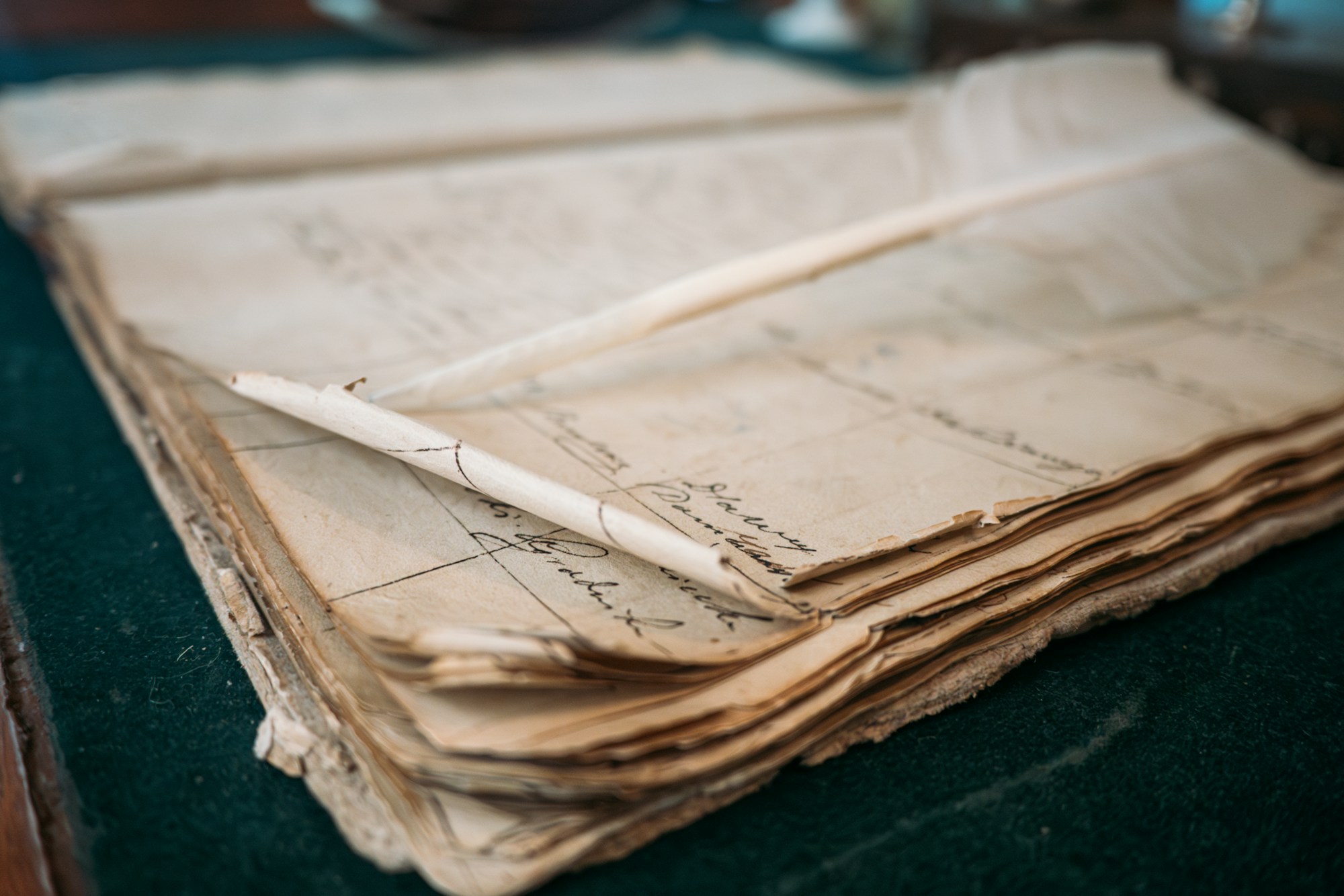Stack of aged handwritten ledger sheets with ruled columns and a quill pen resting on top, on a green cloth